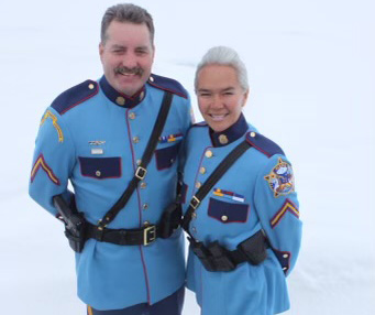 A man and woman pose for a portrait together in their blue Alaska State Troopers dress uniforms.