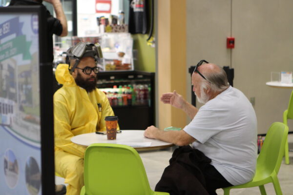 A person in a yellow hazmat suit and facemask sits at a tale with a white man in a white tshirt 