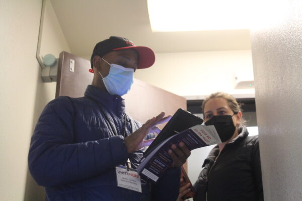 A man with a surgical mask and a baseball hat anad a lanyard scrolls through an ipad in a hallway next to a woman in a black mask