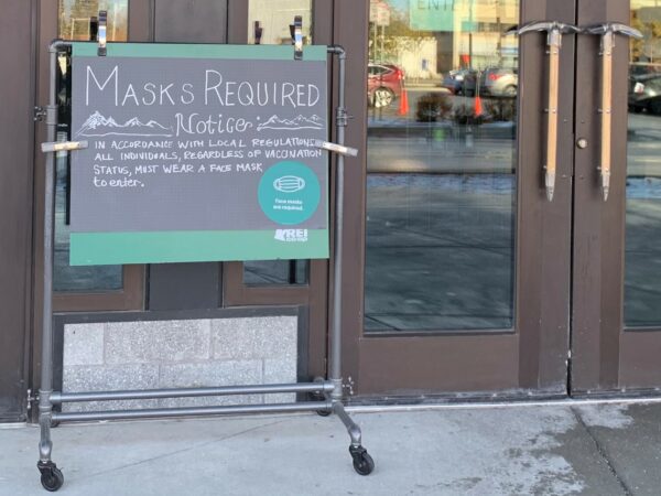A sign outside of a series of doors says "Maska Required Notice: In accordance with local regulations, all individuals regardless of vaccination status, must wear a face mask to enter."