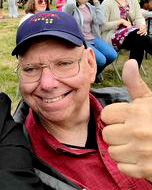 A white man with a baseball hat fives a thumbs up at a field
