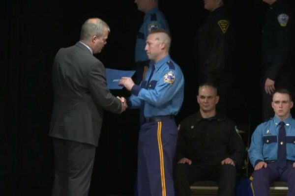 A man in a troopers uniform shakes another man's hand while getting a diploma.