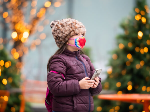 A child in a winter hat, jacket and a mask stands in front of trees with lights on them.