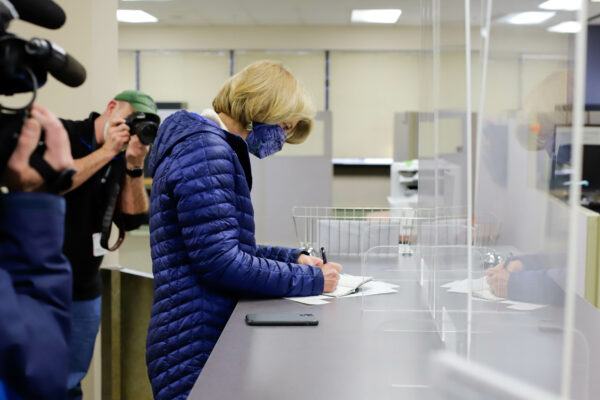 A woman in a blue puffy jacket fills out paperwork at a desk while photojournalists with cameras shoot video and still photos on their cameras.