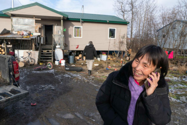 A woman talks on a cell phone outside of a beige house.