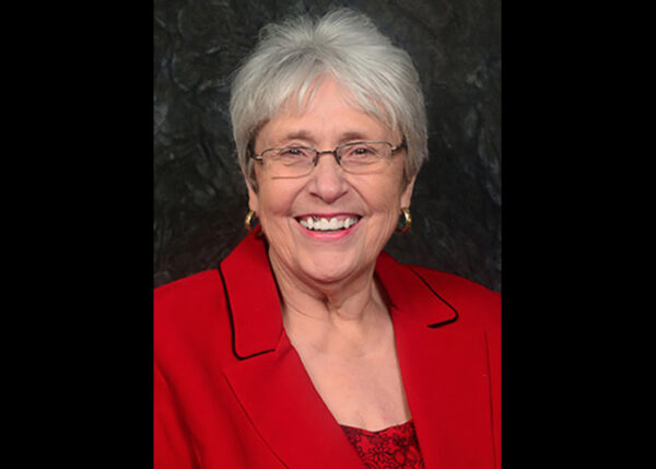 A woman with glasses and gray hair in a red blazer jacket poses, facing the camera and smiling.