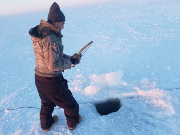 A man in winter clothing stands on ice near a hole with a stick that he's using to ice fish.
