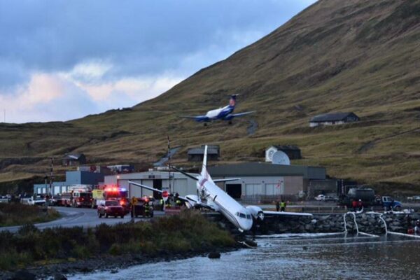 A plane hangs off a runway over water.
