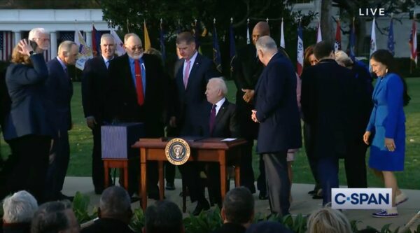 A screenshot of a live video shows people standing around the president who is sitting at a desk outside.