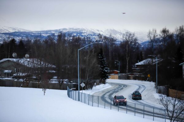 snow covered park, cars driving on a bend in the road, and a plane flies in front of snowy mountains