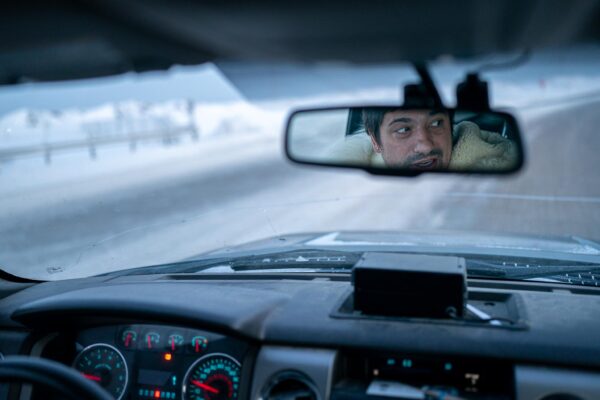 Inside a car, a driver's face can be seen in the rearview mirror.