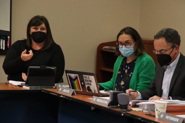 A person in a black blouse and mask sits at a table next to two other people in masks. 