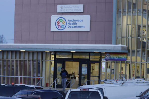 A person leaves the sliding glass doors ofa pink building witha white sign above that says "Anchorage Health Department