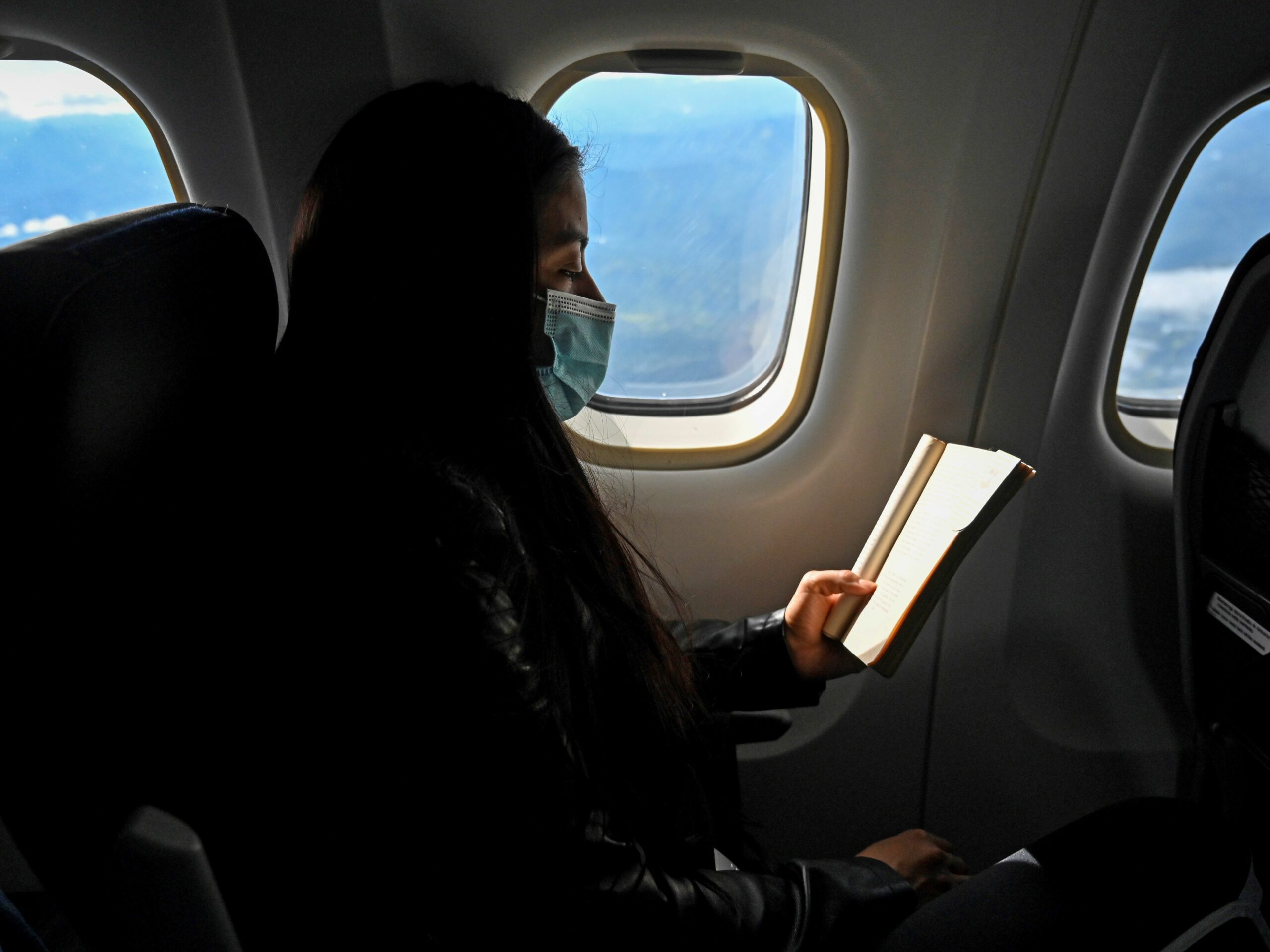 a woman reads a book on a plane while wearing a mask.