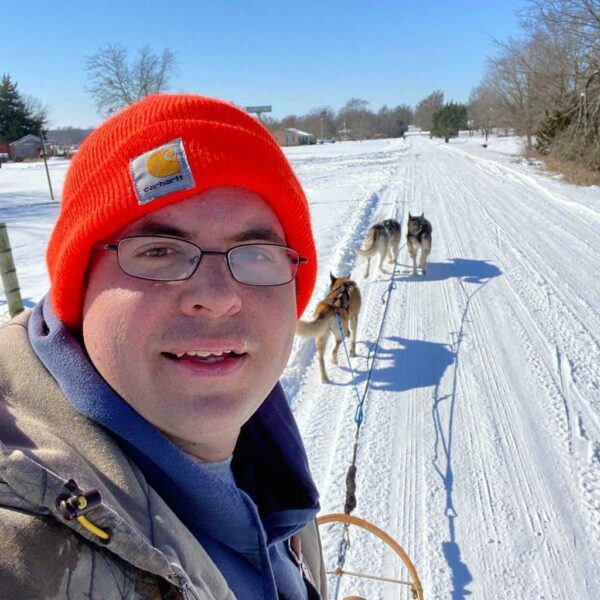 A man in a bright orange carhartt beanie takes a selfie while on a sled being pulled by three dogs