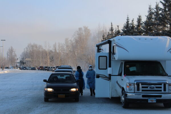 A white camper next to a line of cars in a snowy parking lot with two people in blue hospital gowns walking between them
