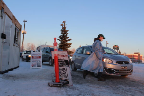 A person in a hospital gown walks around a parked car in a snowy parking lot