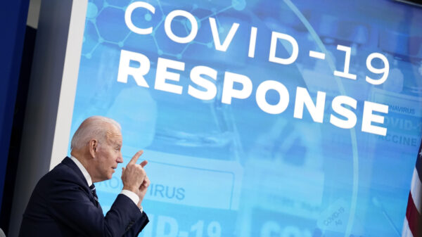 An old white man with white hair in a suit seen in profile in front of a blue projection that says "covid-19 response'