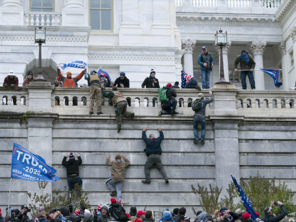 People climb up a wall on a white colonial bilding as a group of people waving flags stands below
