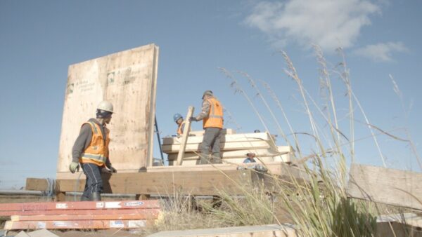 Construction workers stand on a wooden platform with a stack of lumber next to them on a sunny day in the tundra