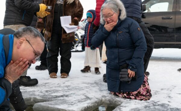 A man and woman wipe their face with water, kneeling on river ice.