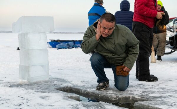 A man wipes his face with water, kneeling in front of a cross cut into river ice.