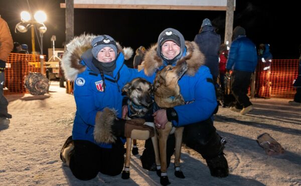 Two people in big winter coats pose with two sled dogs