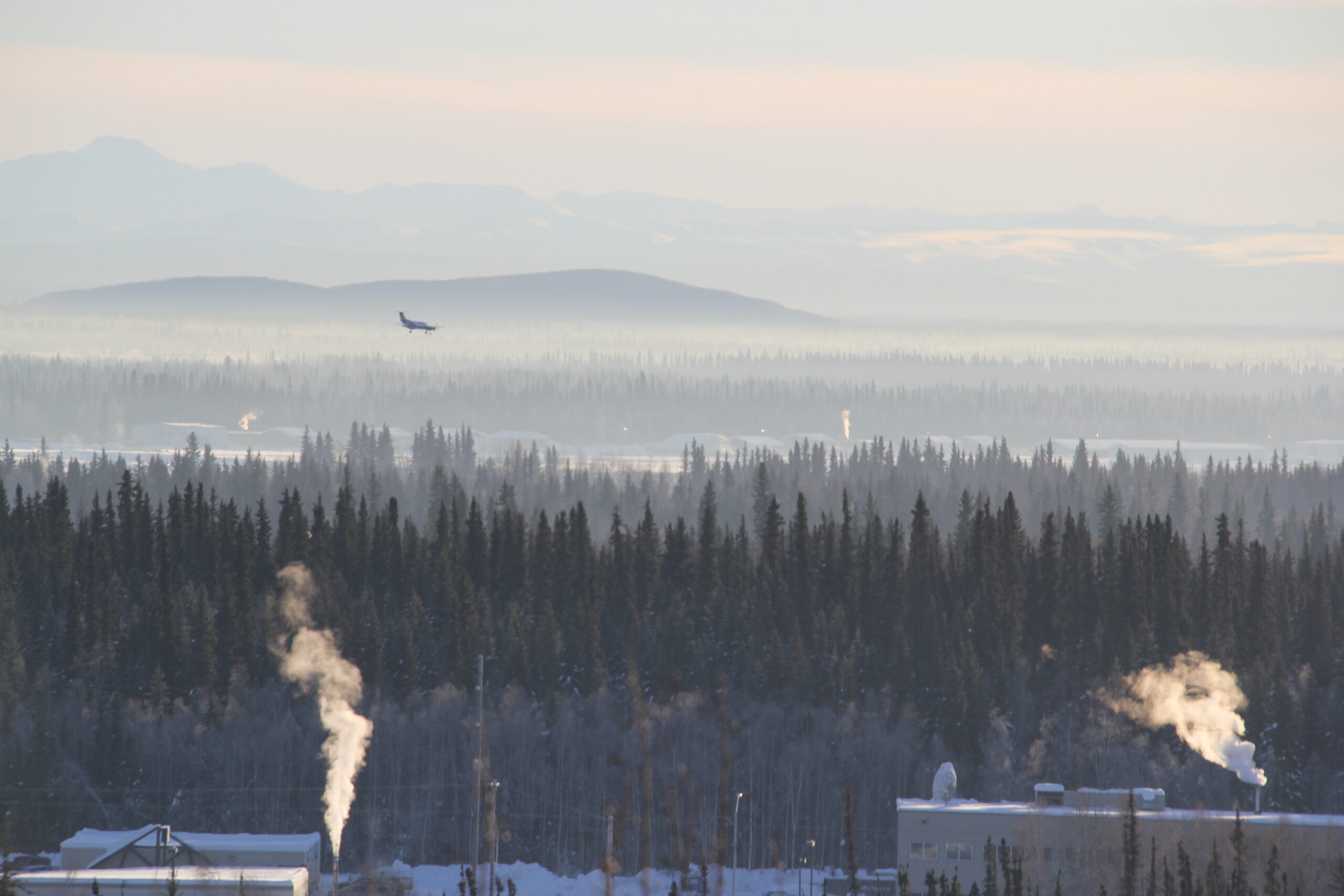 Smoke and haze rising above mostly spruce as a plane lands in the distance