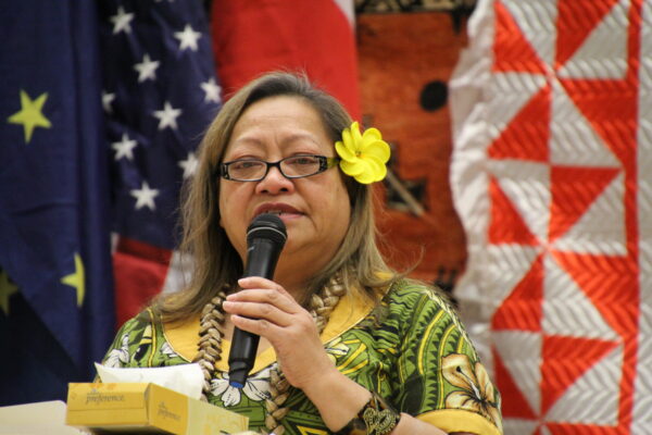 A woman in a green floral shit, glasses and a yellow flower in her hair, speaks into a microphone. 