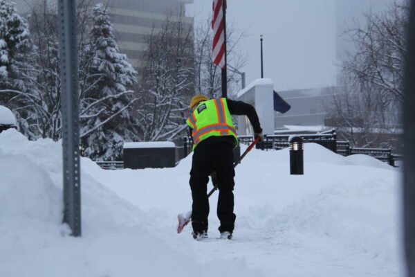 A man in a yellow safety vest shovels snow in front of an american flag