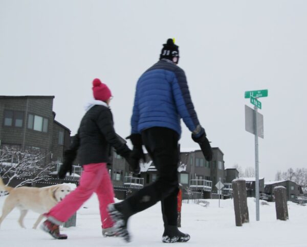 Two people in winter gear and a dog walk along a snowy trail by some houses