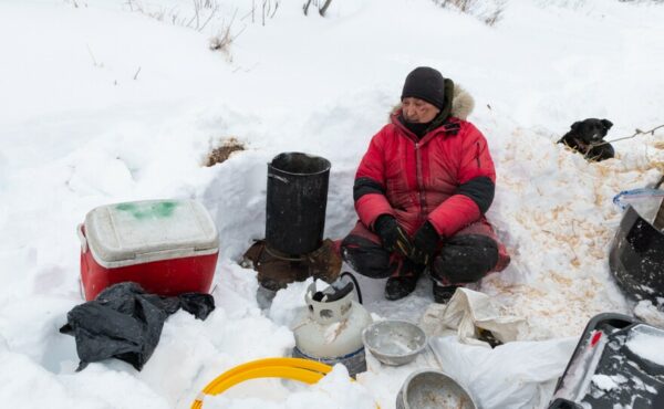 A musher sits outside near a stove in the snow