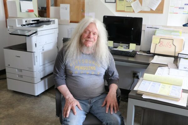 A man with long white hair and a white beard seated in an office