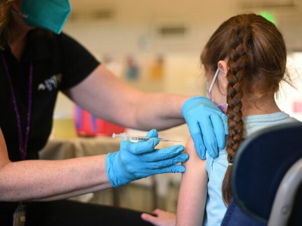 Seen from behind , a girl with a braid sits in a chair while an adult in a black shirt gives an injection in her shoulder wearing blue gloves
