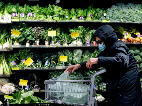 A person wearing a black parka and a mask pushing a shopping cart through a produce aisle