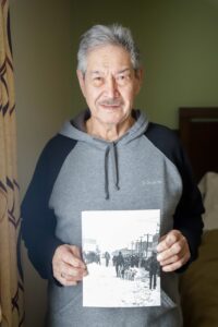 Portrait of a man in a gray hoodie holding an old photo of a dog team running down Front Street in Nome