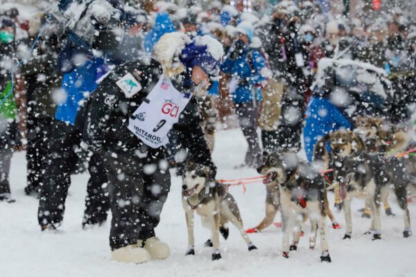 A woman dressed in black snow gear wearing a bib #9 holds a dog that's tied up to a sled with a bunch of people in the background