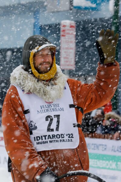 A man in an orange parka waves to the crowd with his hand on a sled runner