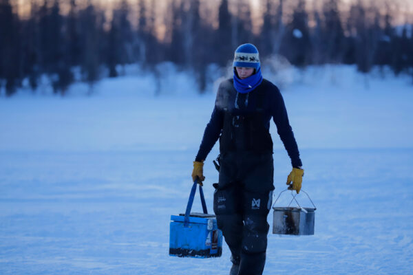 A frosty musher walks in the snow carrying two containers
