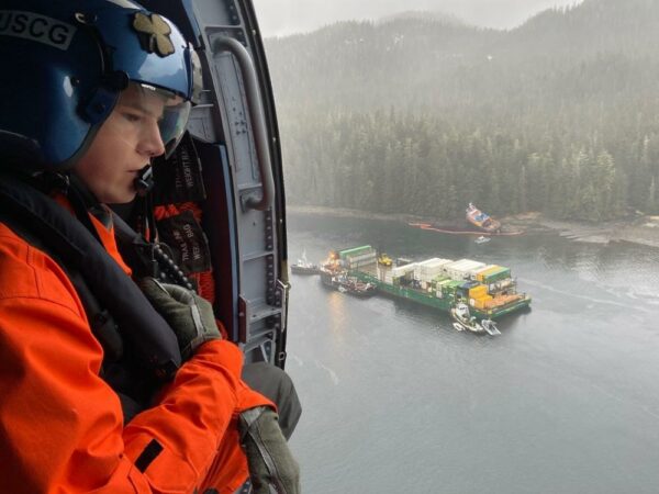 A Coast Guard member looks out the open door of a helicopter at a container barge surrounded by smaller vessels