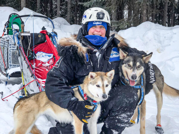 A woman ina helmet and parka holds two dogs around her arms