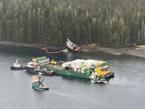 Small vessels surround a container barge as a grounded tugboat lays on the shore