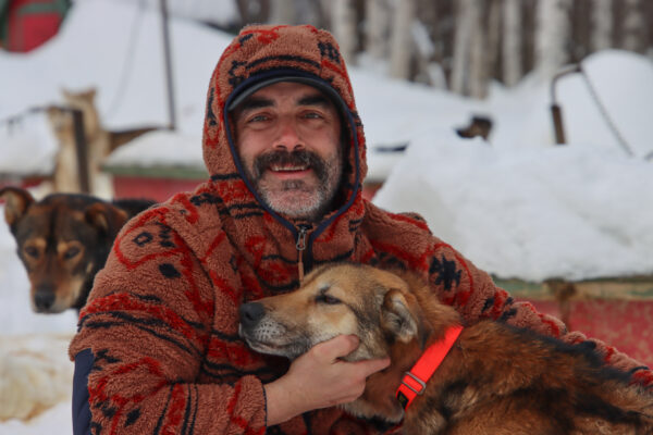 A man in a fuzzy orange hoodie and a mustache kneels next to a dog, petting it