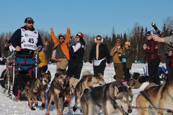 A man mushing a dog team holds a silver beer in his hand as a man cheers berhind him