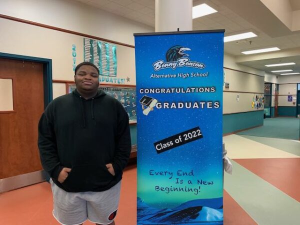 Young Black man smiles slightly while standing next to a blue sign that reads "Benny Benson Alternative High School Congratulations Graduates Class of 2022."