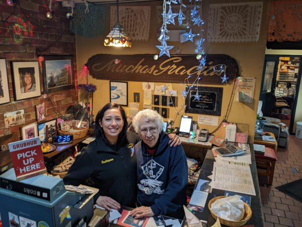 A younger woman and an older woman stand behind a counter surrounded by photos and decorations
