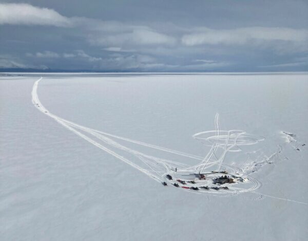 An aerial view of a crowd of people with snowmachines gathered in a remote spot on flat, snowy tundra