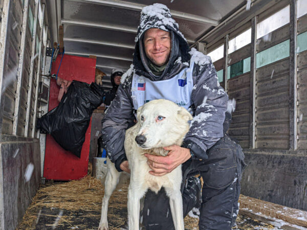 A man in musher's gear kneeling with his arms around a white, blue-eyed sled dog