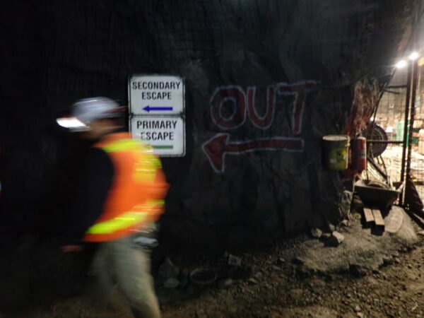 A man in an orange vest walking past  exit signs in a mine