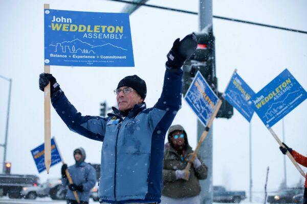 a person waves while holding a sign that says "John Weddleton - Assembly - balance, experience, commitment"on a snowy day. Others in the background wave the same sign